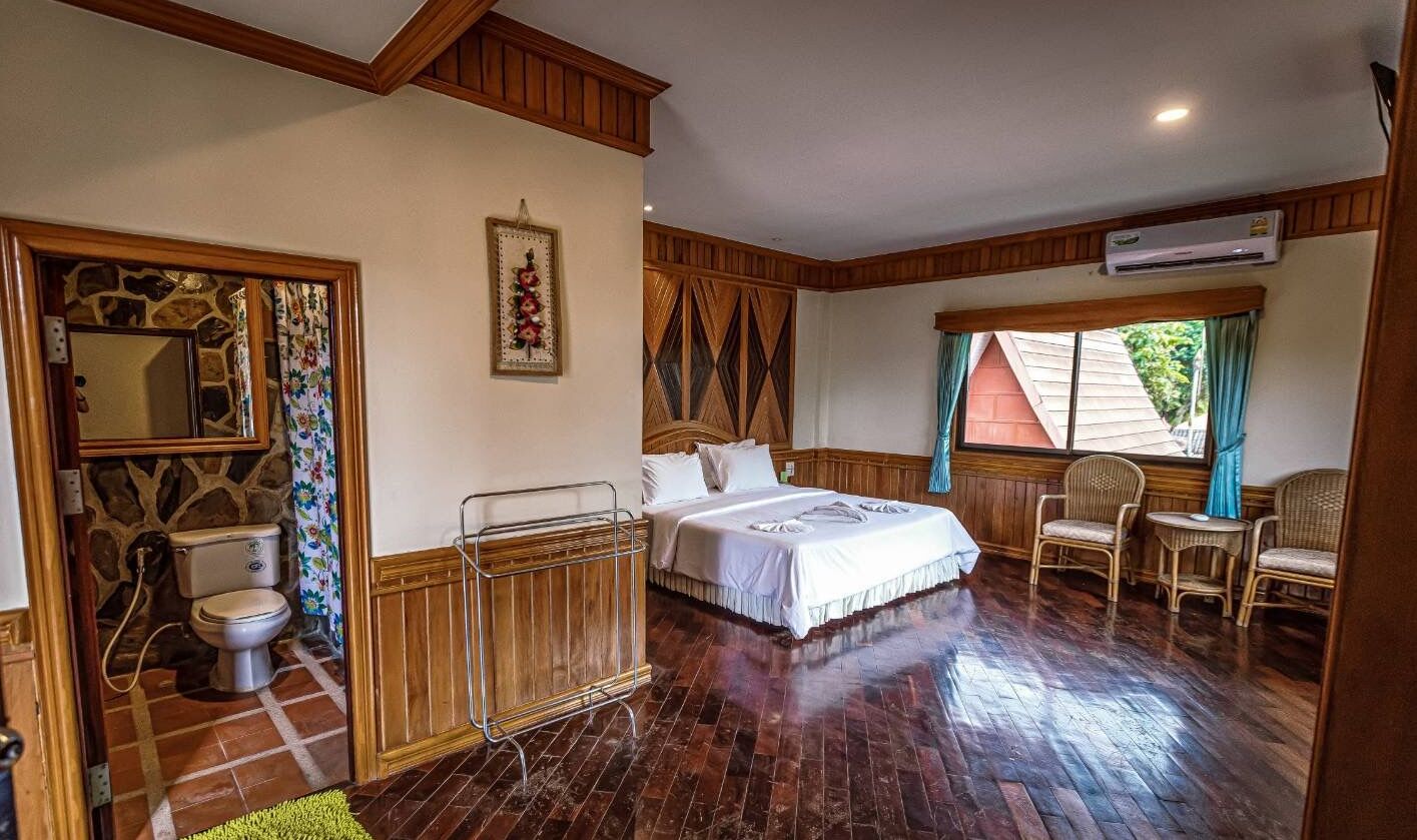 A bedroom with a bed, a chair, a small table, a window with curtains, a wall-mounted air conditioner, and a wooden floor.