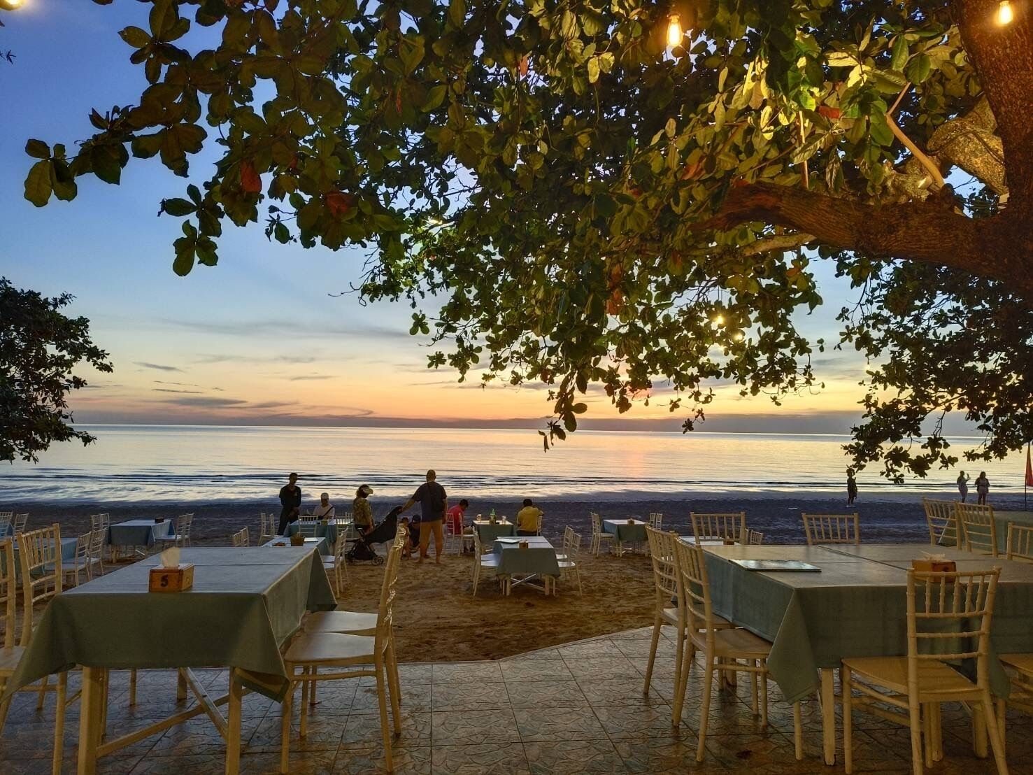 Een buitenterras met tafels en stoelen, uitkijkend op een strand en de oceaan bij zonsondergang.