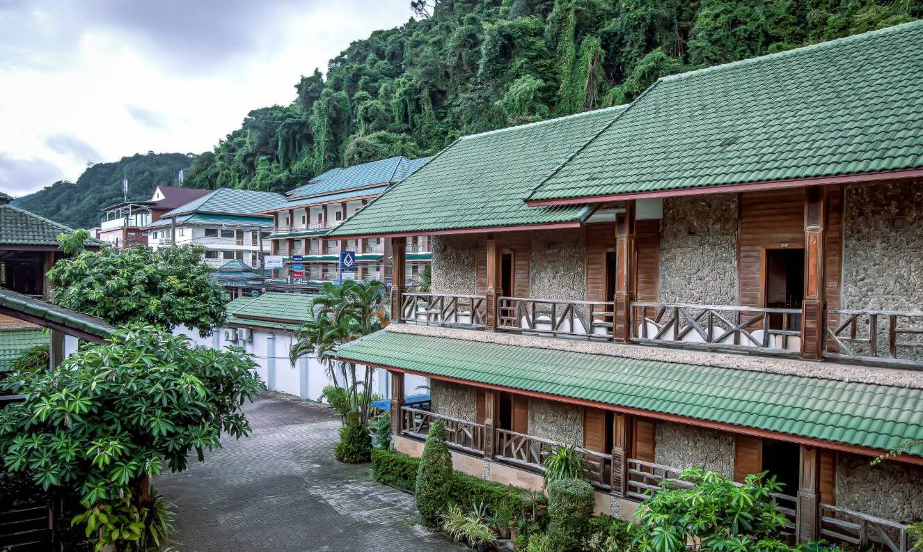 A row of traditional-style buildings with green roofs and wooden balconies, surrounded by lush greenery and hills.