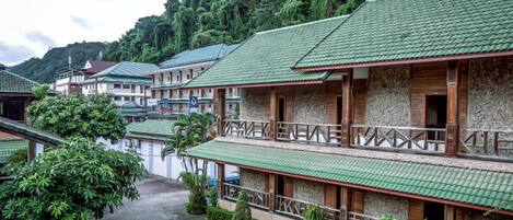 A row of traditional-style buildings with green roofs and wooden balconies, surrounded by lush greenery and hills.