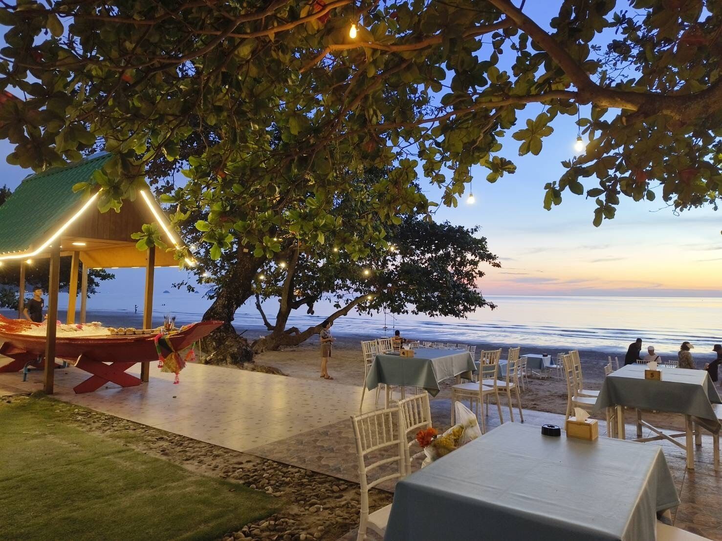 Un área de comedor al aire libre con mesas y sillas bajo un árbol, con vistas a la playa al atardecer.