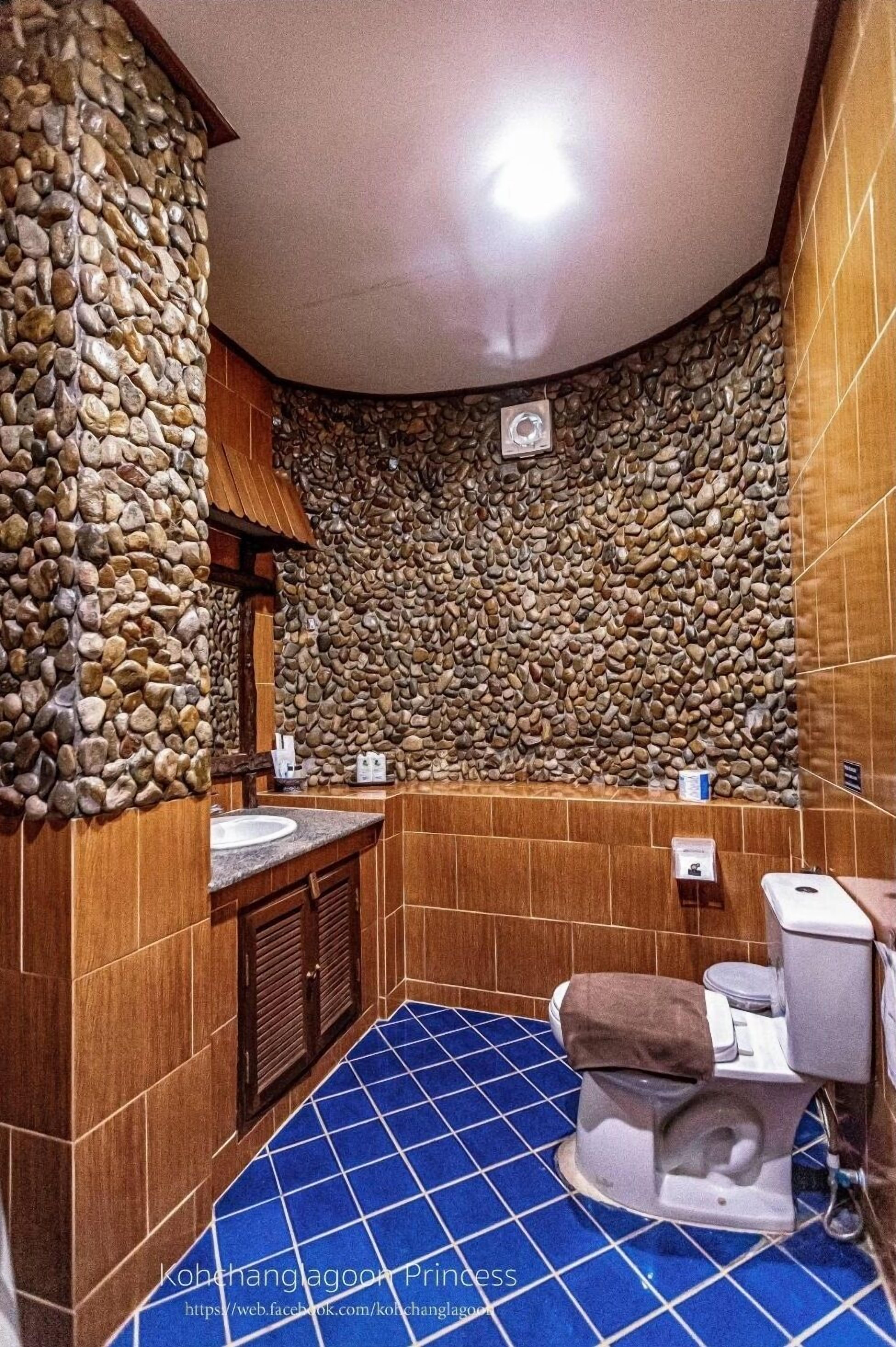 A bathroom with a stone wall, wooden cabinets, a blue-tiled floor, and a toilet.