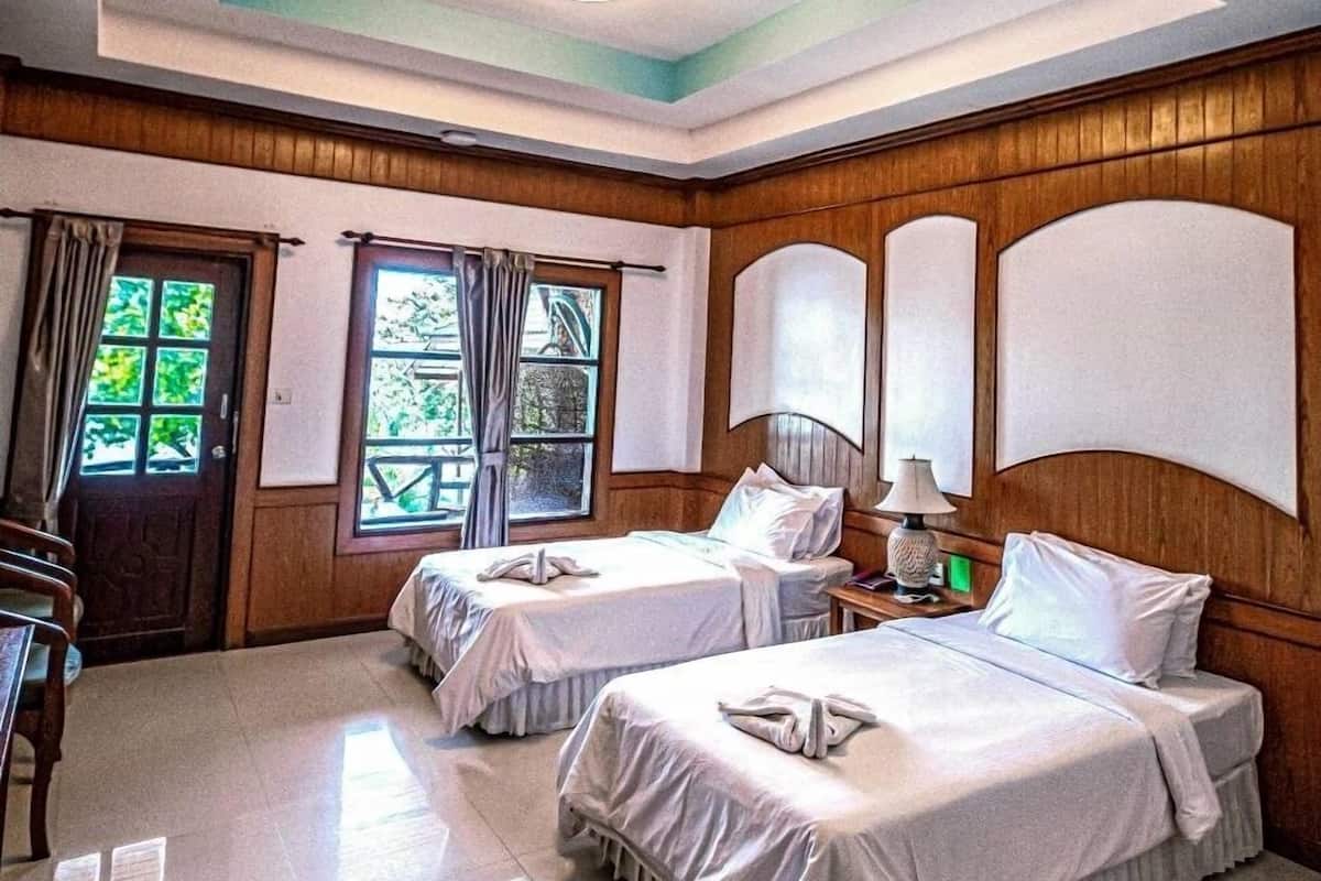 A hotel room with two beds, wooden paneling, a ceiling light, and a view of greenery outside.