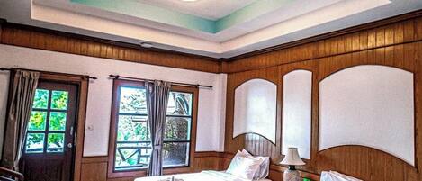 A hotel room with two beds, wooden paneling, a ceiling light, and a view of greenery outside.