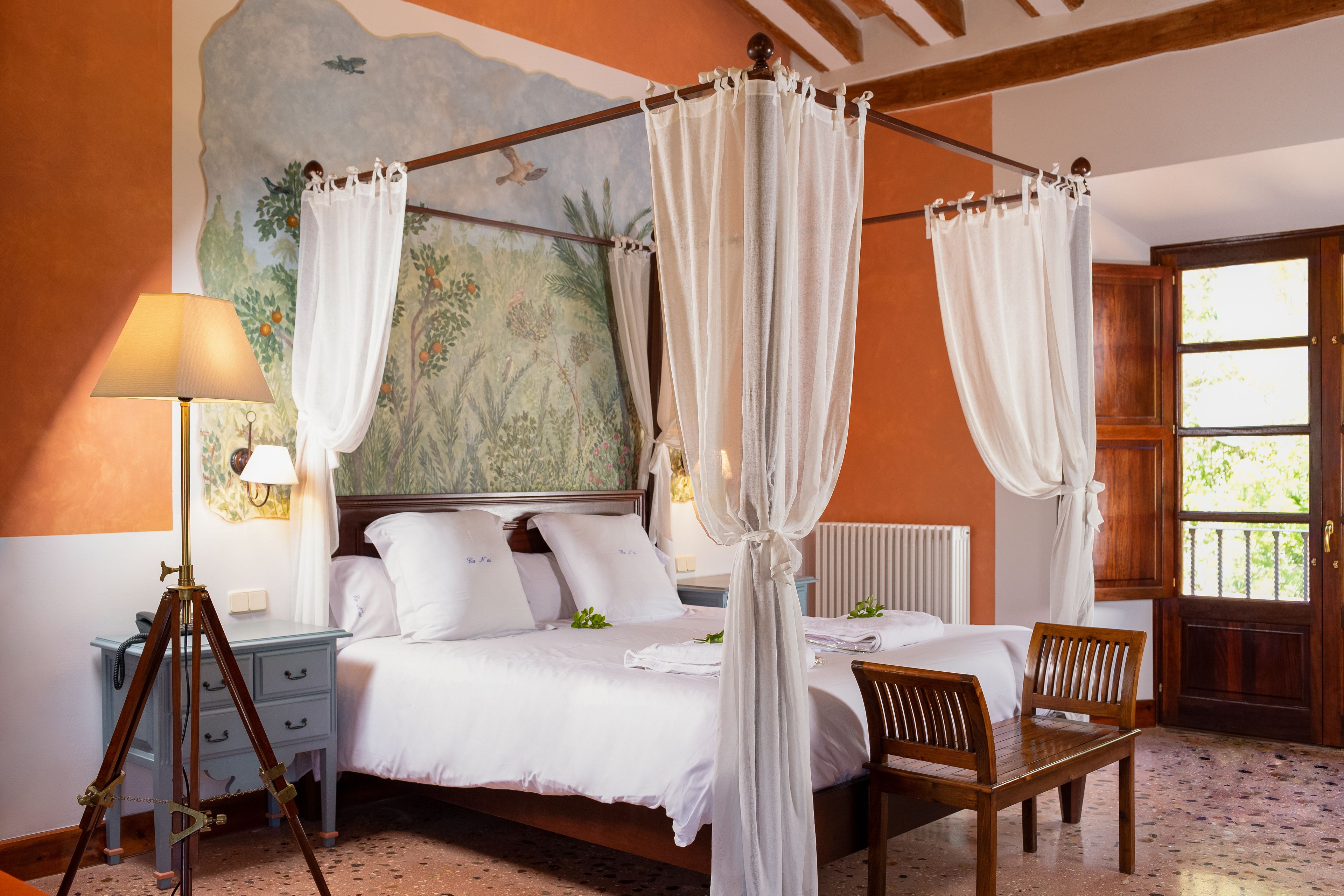 A bedroom with a four-poster bed, a bedside table with a lamp, a chair, and a mural on the wall.