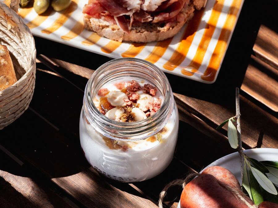 A table with a variety of food items including bread, cheese, olives, and a jar of yogurt with fruit.