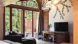 A bedroom with a bed, a desk, a chair, and a TV. There is a window with a view of trees and a balcony.