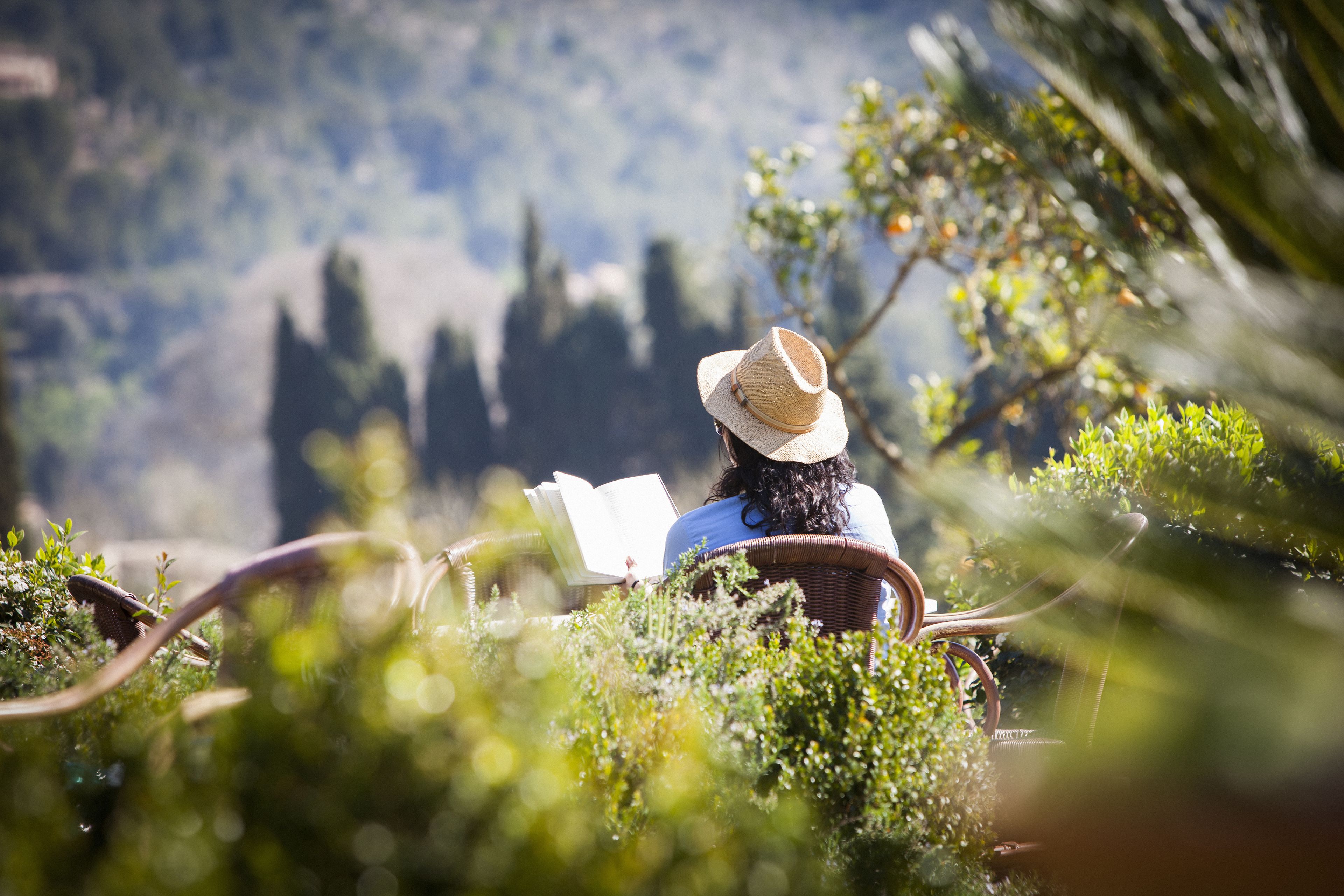 Una persona sentada en una silla de mimbre leyendo un libro, rodeada de follaje y con vistas a un paisaje montañoso.