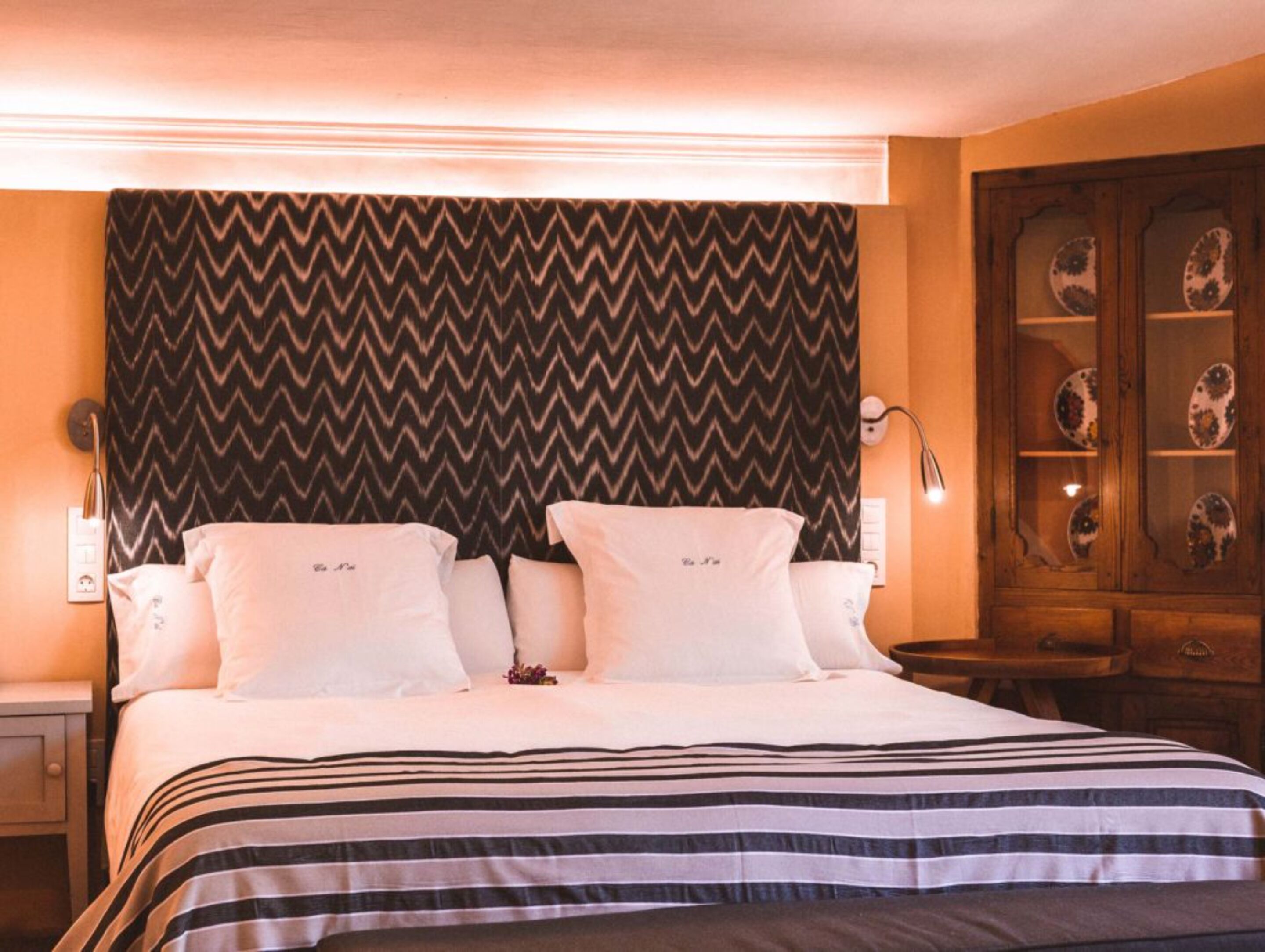 Una habitación de hotel con una cama, dos mesitas de noche, una lámpara y un armario de madera con platos decorativos.
