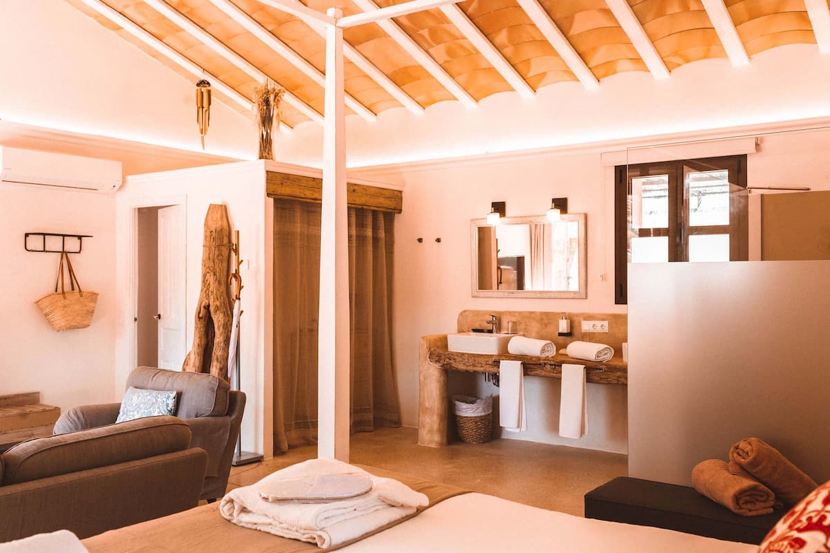Een kamer met een bed, een massagestoel, een houten vloer, een badkamer met wastafel en spiegel, en een plafond met houten balken.