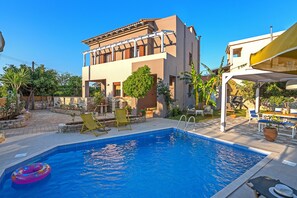 Una piscina con agua cristalina azul, rodeada por un patio con sillas de descanso y una zona de estar cubierta.