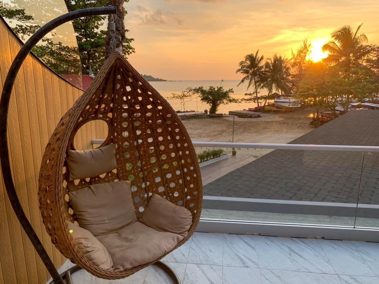 A hanging chair with cushions on a balcony overlooking a beach with palm trees and a sunset.