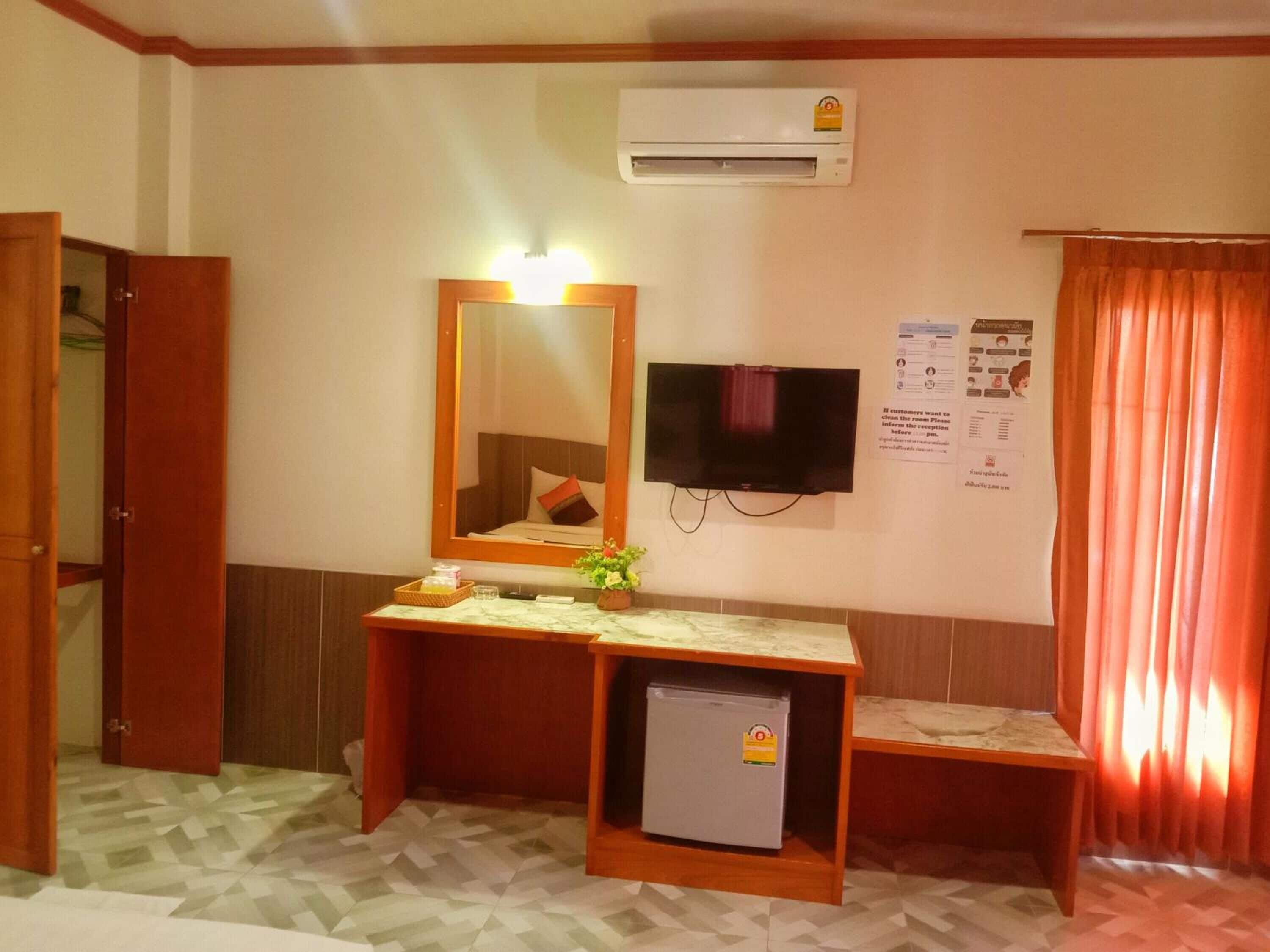 A hotel room with a wooden desk, a small refrigerator, a TV mounted on the wall, and a mirror above the desk.