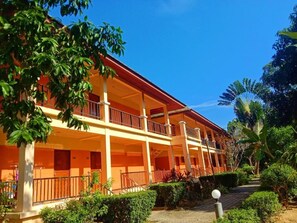 Um edifício de dois andares com varandas, cercado por vegetação e um céu azul límpido.