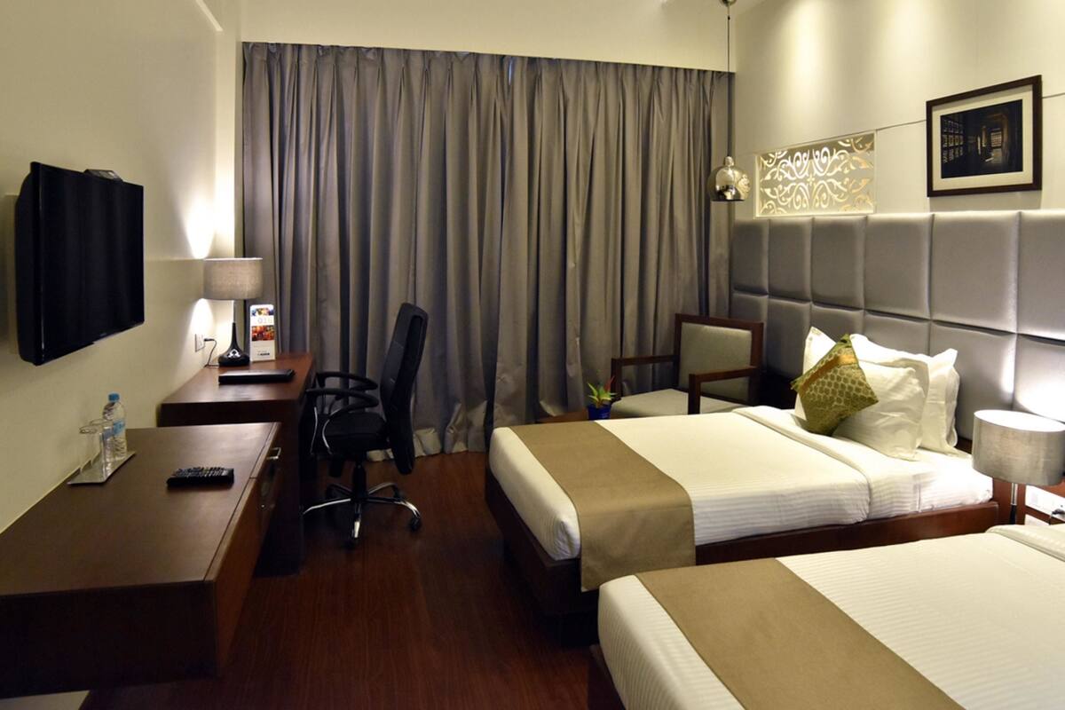A hotel room with two beds, a desk, a chair, a TV, and a window with curtains.