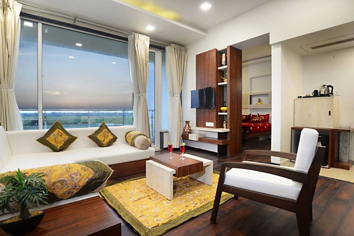 A modern living room with a sofa, armchair, coffee table, and a TV. There is a view of the outdoors through large windows.