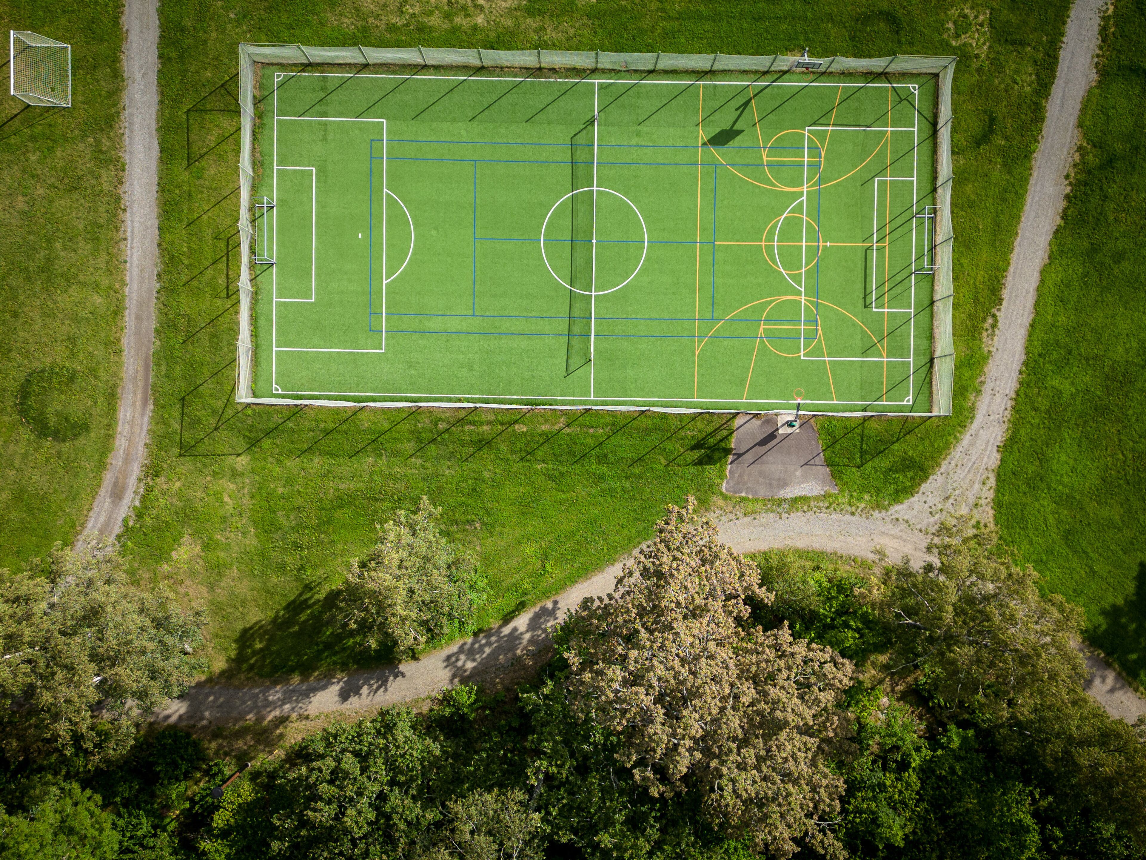 An aerial view of a sports field with marked lines for soccer and basketball, surrounded by trees and a dirt path.