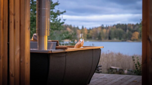 A wooden deck with a grill, a glass of white wine, and a view of a lake and forest. - Marholmen Stugor (Norrtälje)