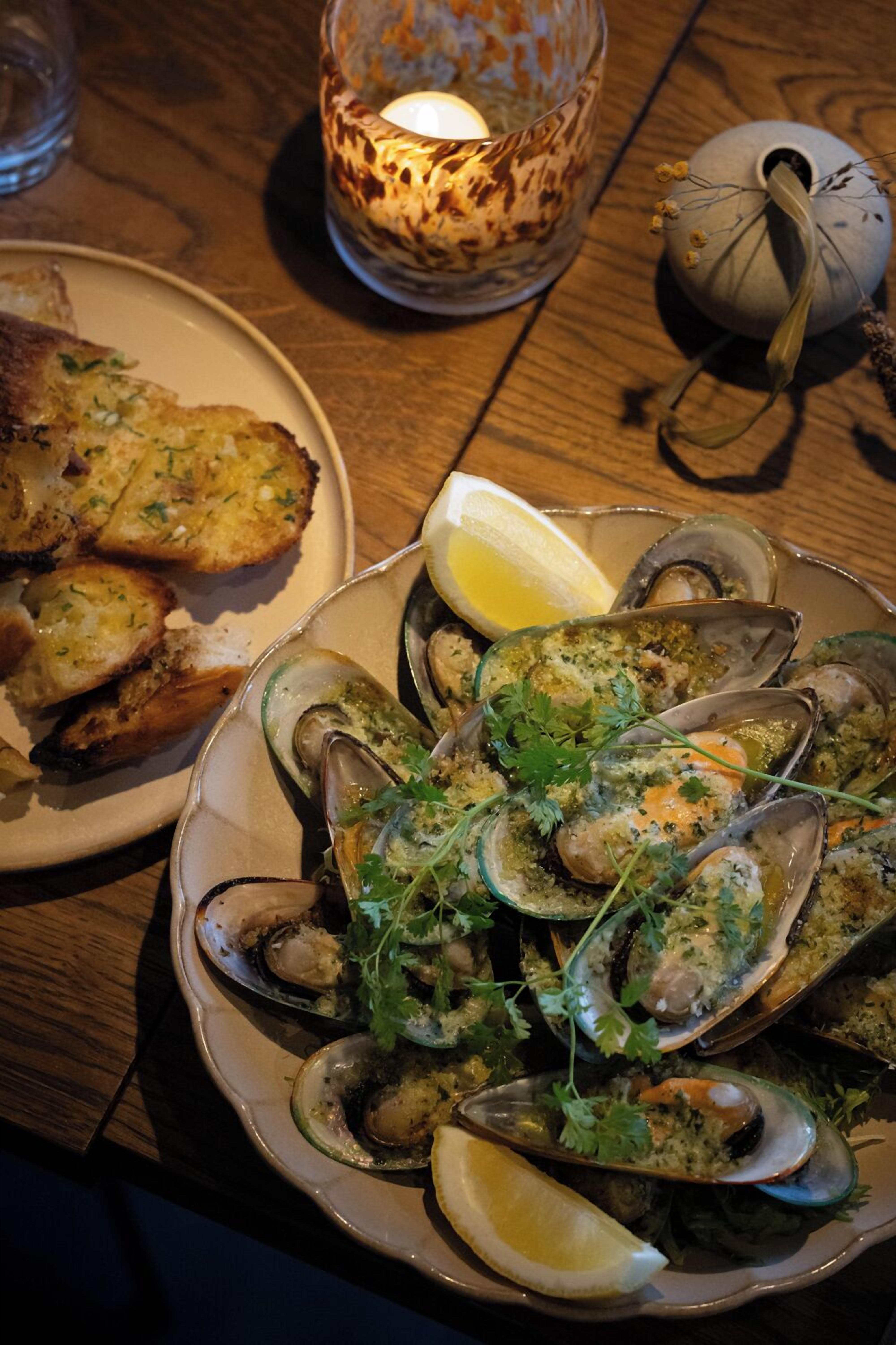 A plate of cooked mussels garnished with herbs and lemon slices, accompanied by toasted bread, on a wooden table.