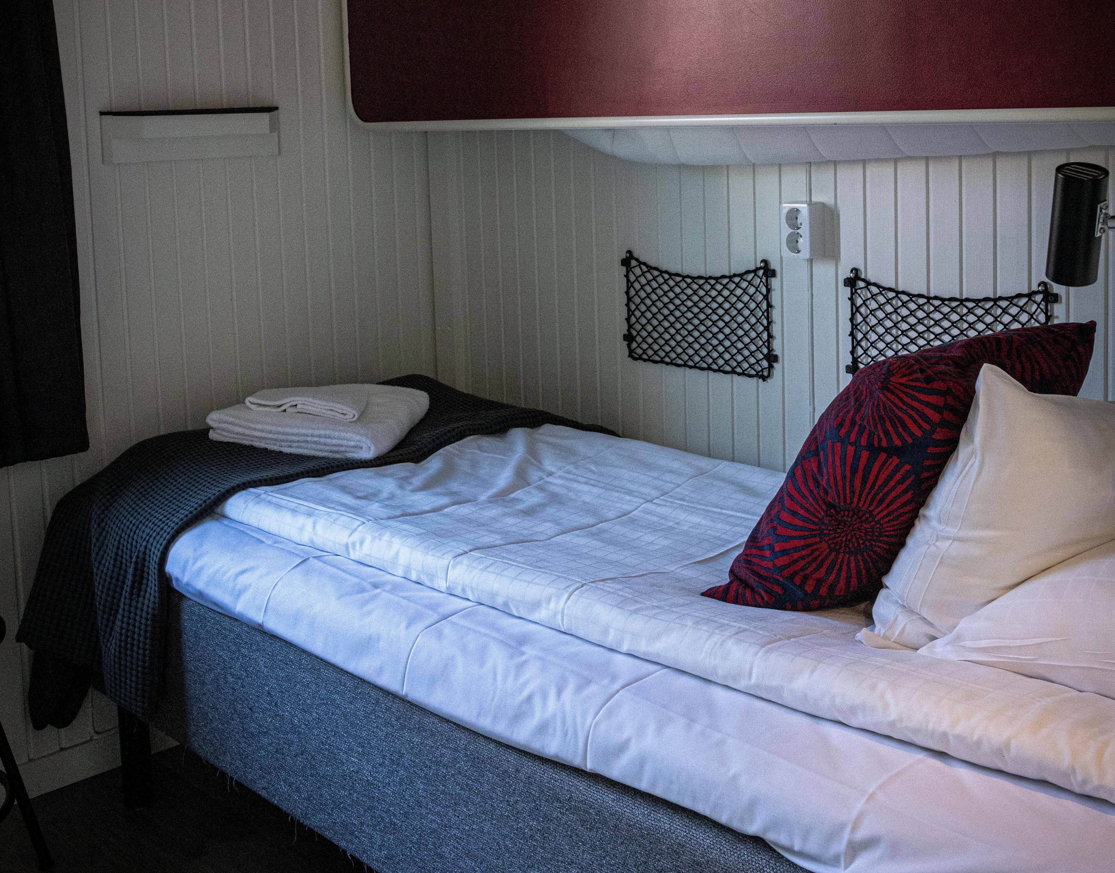 A neatly made bed with a blue and white duvet, a folded white towel, and a red and black patterned pillow. There is a black headboard with a net design, and a wall-mounted light fixture above the bed.