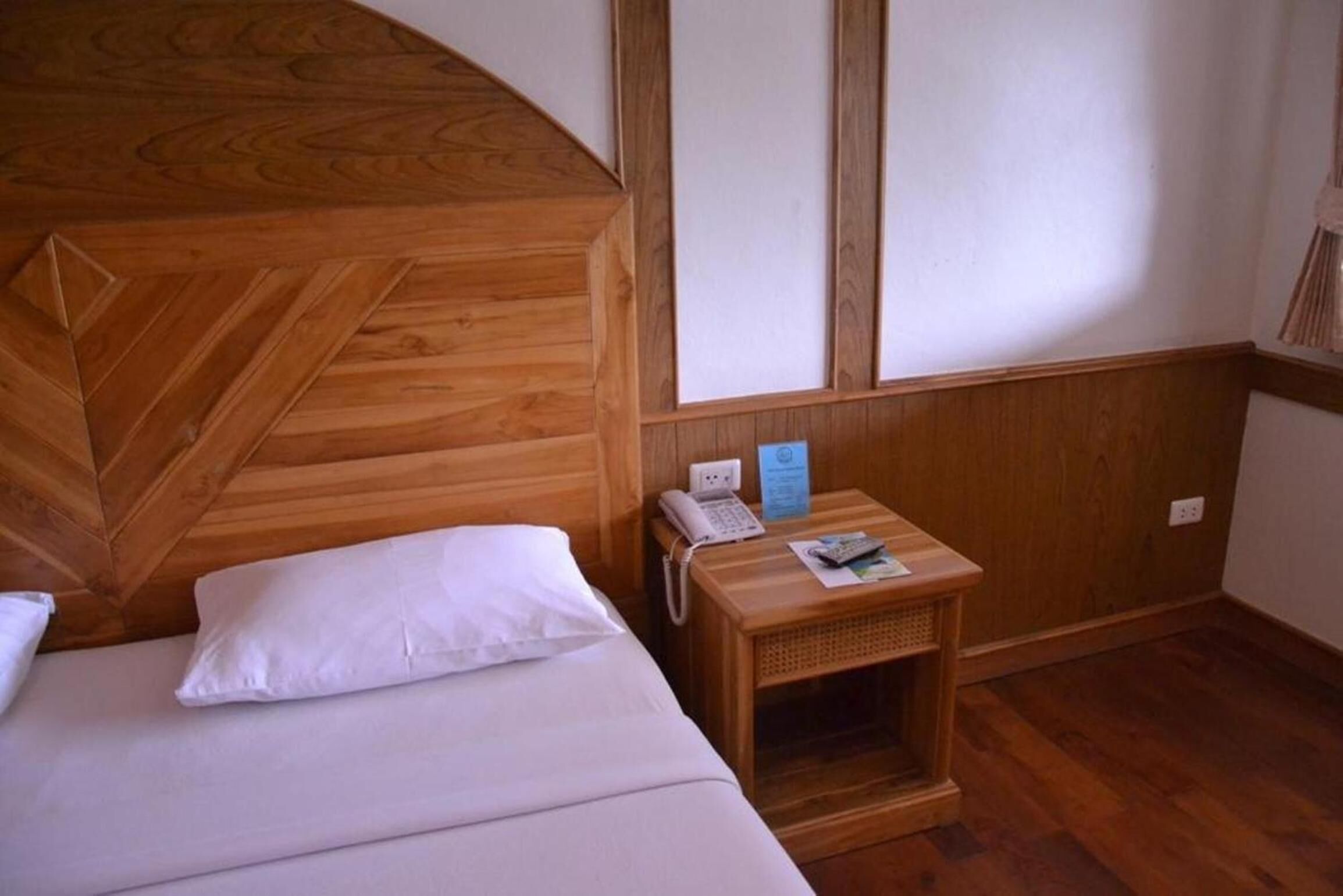 A wooden bed with a white pillow and a small bedside table with a phone and a brochure.
