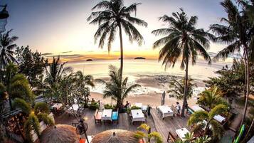 A beachside restaurant with outdoor seating overlooking the ocean, palm trees, and a sunset view.