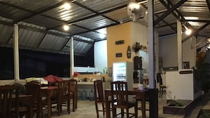 Un comedor interior con sillas y mesas de madera, una zona de cocina con refrigerador y pileta, y un mostrador de recepción.