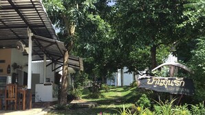 Zona de recepción con un cartel, un espacio de descanso cubierto y un jardín con flores amarillas.