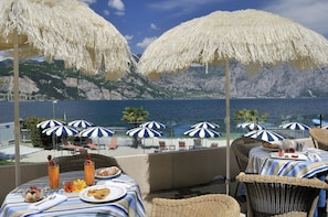 A seaside restaurant with tables and chairs, striped tablecloths, and white thatched umbrellas. - Hotel Castello Lake Front (Malcesine)