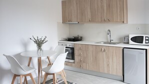 A modern kitchen with wooden cabinets, a white countertop, a built-in oven, a sink, a microwave, and a dishwasher. There is a round dining table with two white chairs and a vase with dried flowers. - Axos Apartments (Hersonissos)