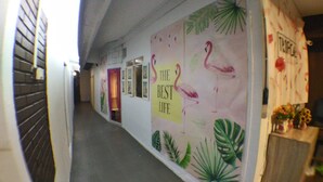 Un pasillo con un mural de flamencos y hojas tropicales, una puerta con cortina rosa y una pared con un letrero que dice 'TROPICAL'.
