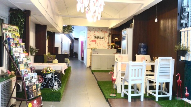 Un pasillo estrecho con una mesa de recepción, sillas blancas y una pequeña mesa. A la izquierda, hay libros y flores, y a la derecha, un dispensador de agua.