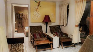 Una habitación con dos sillas de madera, una lámpara roja, un cuadro de flores y una ventana con cortinas.