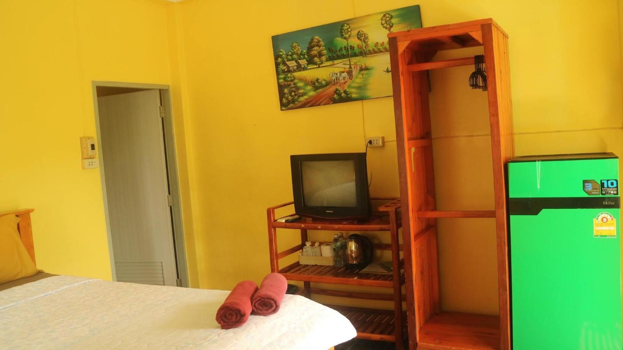 Una habitación con una cama, un televisor sobre un soporte de madera, un refrigerador verde y un cuadro en la pared.