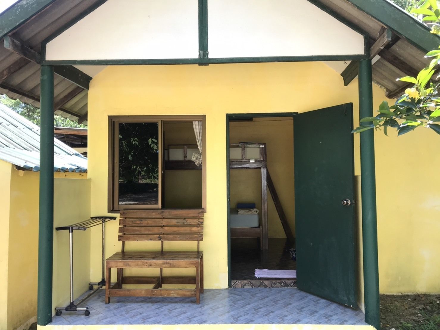 Un petit bâtiment jaune, avec un banc en bois à l’extérieur, un chariot en métal et une porte donnant sur une pièce où se trouvent un lit et une petite table.