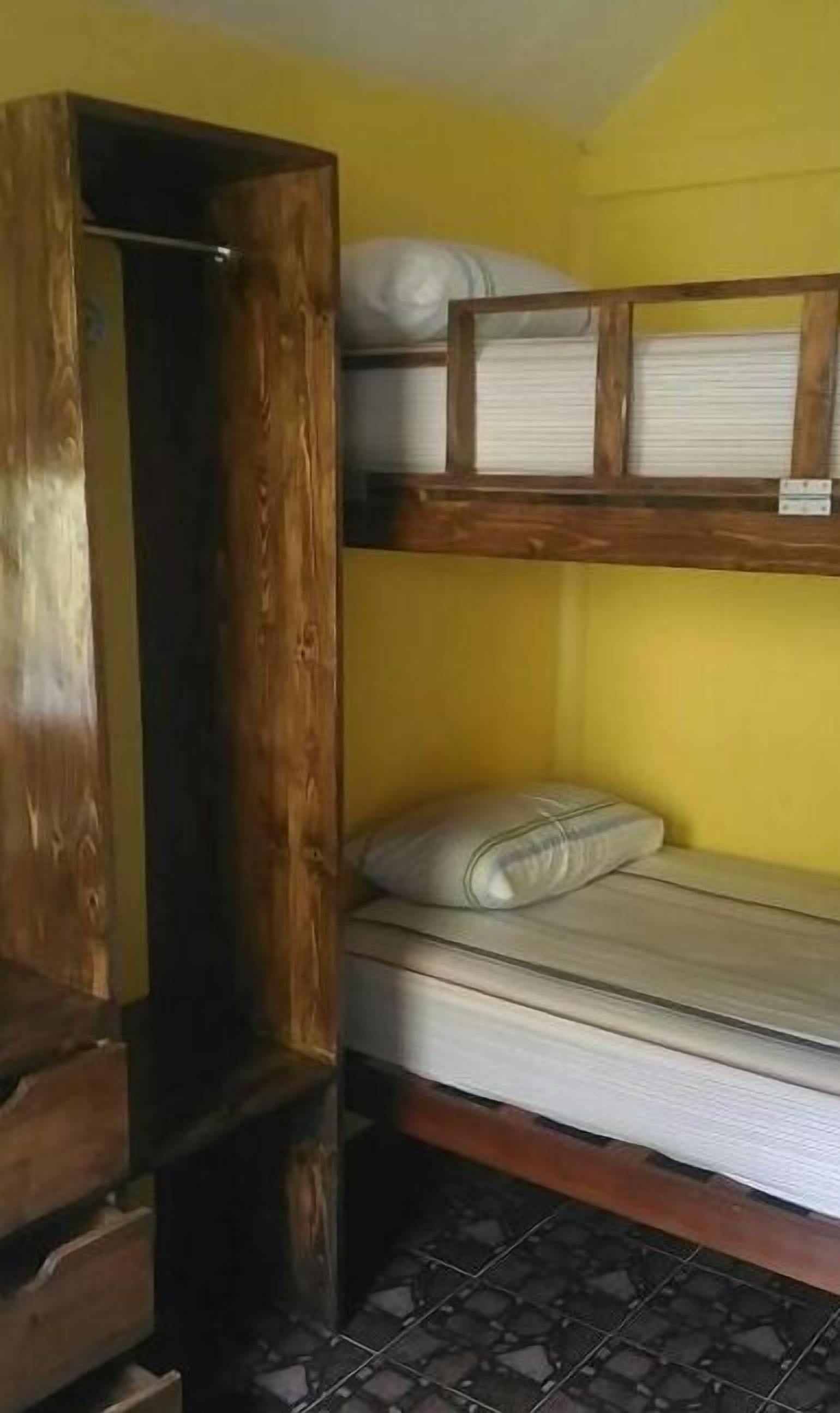 Un lit superposé avec une structure en bois et un matelas, une armoire en bois et un sol carrelé.