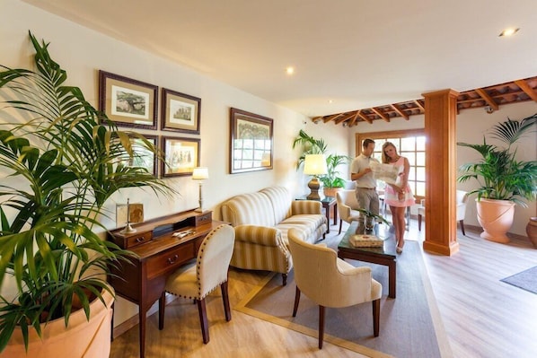 Un salon spacieux comprenant un canapé, des fauteuils, une table basse et un bureau. Les murs sont ornés de tableaux encadrés, il y a une grande plante et une fenêtre laissant entrer la lumière naturelle.