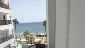 A balcony with a round table and two chairs, overlooking a beach with palm trees and umbrellas. - Palm Sea Hotel Apartments 2 (Larnaca)