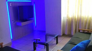 A room with a TV mounted on a wall, a small table, a sofa, and a window with curtains. - Palm Sea Hotel Apartments 2 (Larnaca)