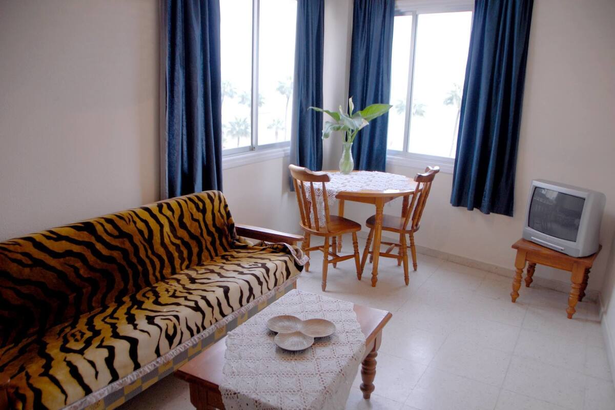 A room with a tiger-patterned sofa, a small table with a lace tablecloth, a wooden chair, a television on a stand, and a window with blue curtains.