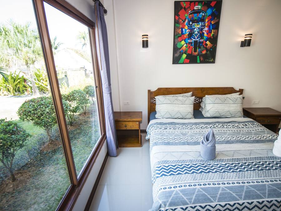 Un dormitorio con un ventanal grande, una cama con ropa de cama estampada, dos mesitas de noche y una obra de arte colorida en la pared.