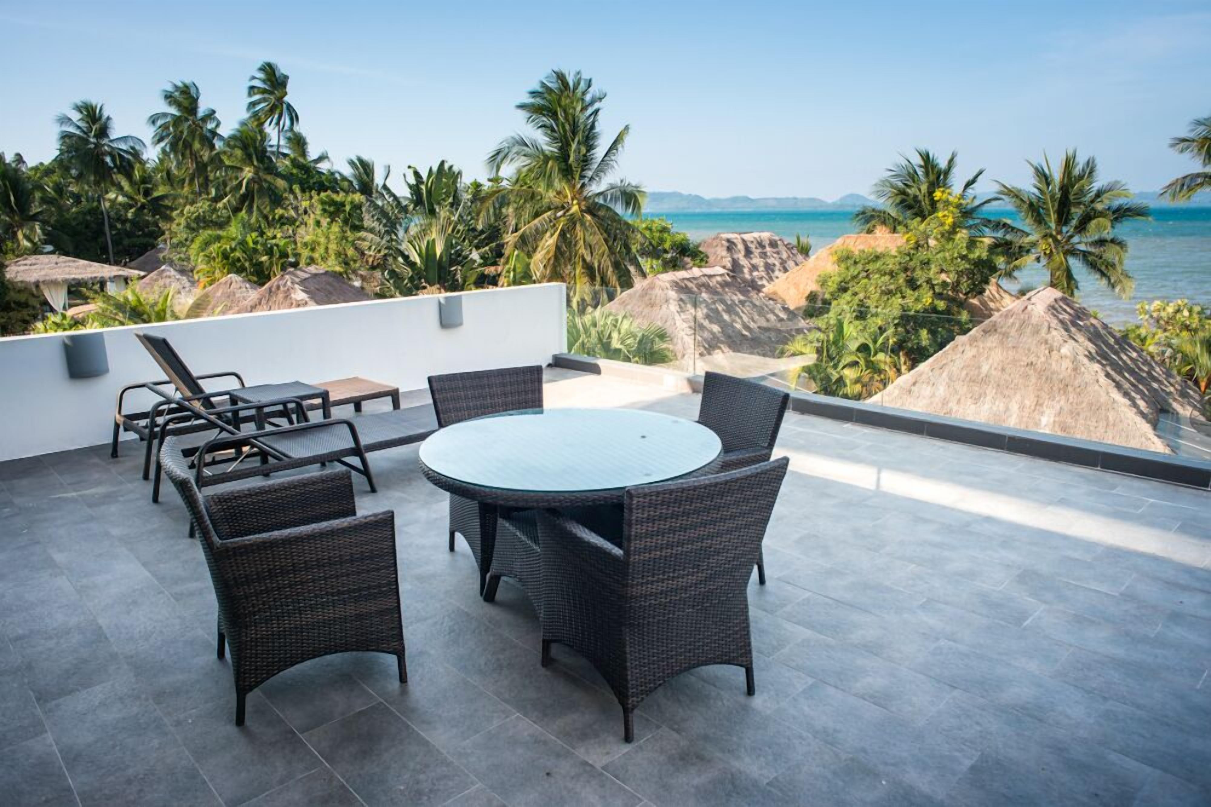 Terraza en la azotea con una mesa redonda y sillas, con vista a una playa con palmeras y chozas de techo de paja.