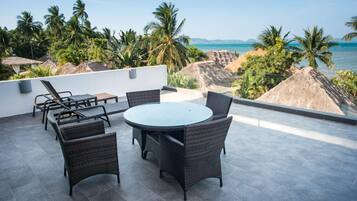 Terraza en la azotea con una mesa redonda y sillas, con vista a una playa con palmeras y chozas de techo de paja.