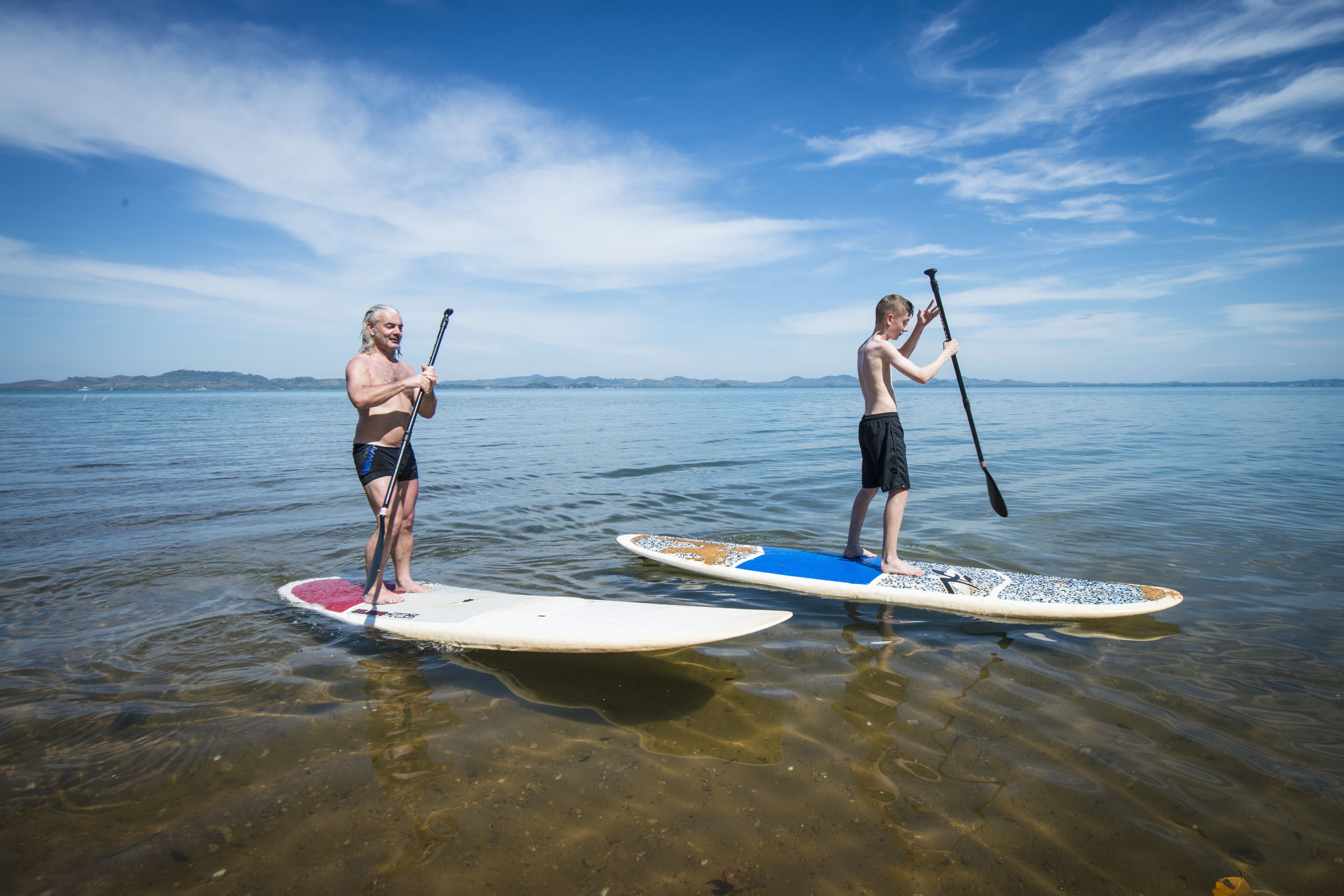 Dos personas practican remo de pie sobre aguas tranquilas bajo cielos despejados.