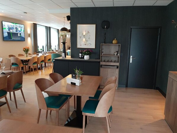 A modern dining area with wooden tables and teal chairs, a reception desk with a framed artwork, and a television mounted on the wall. - Hotel Limburgia (Riemst)