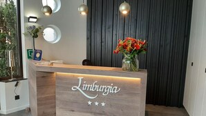 Een receptiebalie met een houten afwerking waarop de naam 'Limurgia' staat. Daarboven hangen drie hanglampen, en op de balie staat een vaas met rode bloemen.