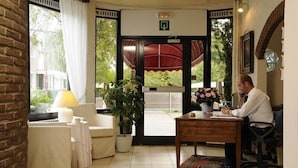 Een hotellobby met een receptiebalie, een zithoek met een bank en een tafel, een kamerplant, en uitzicht naar buiten door een glazen deur.