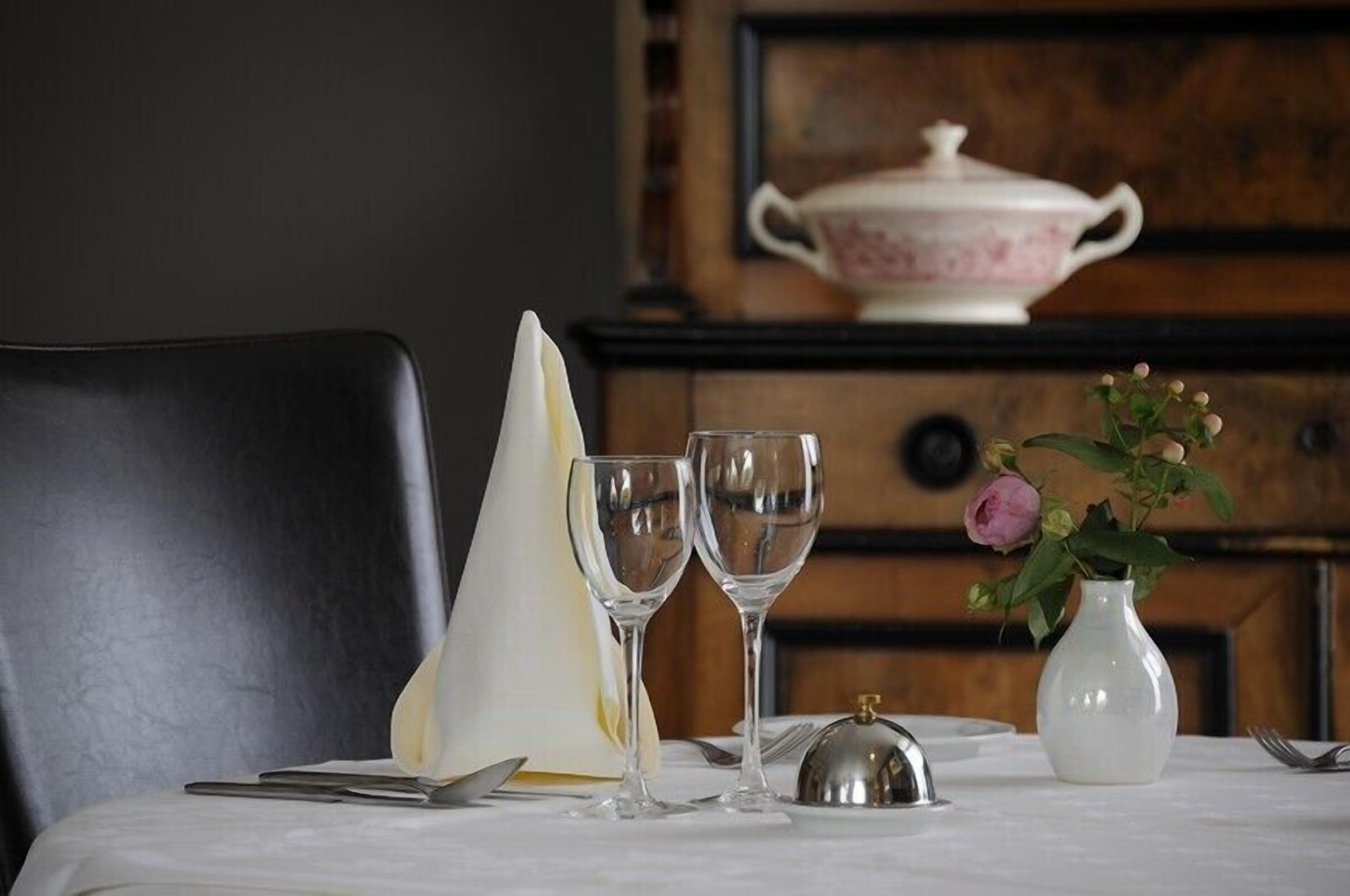 Una mesa de comedor con dos copas de vino, un mantel blanco, una campana de plata y un jarrón blanco con flores.