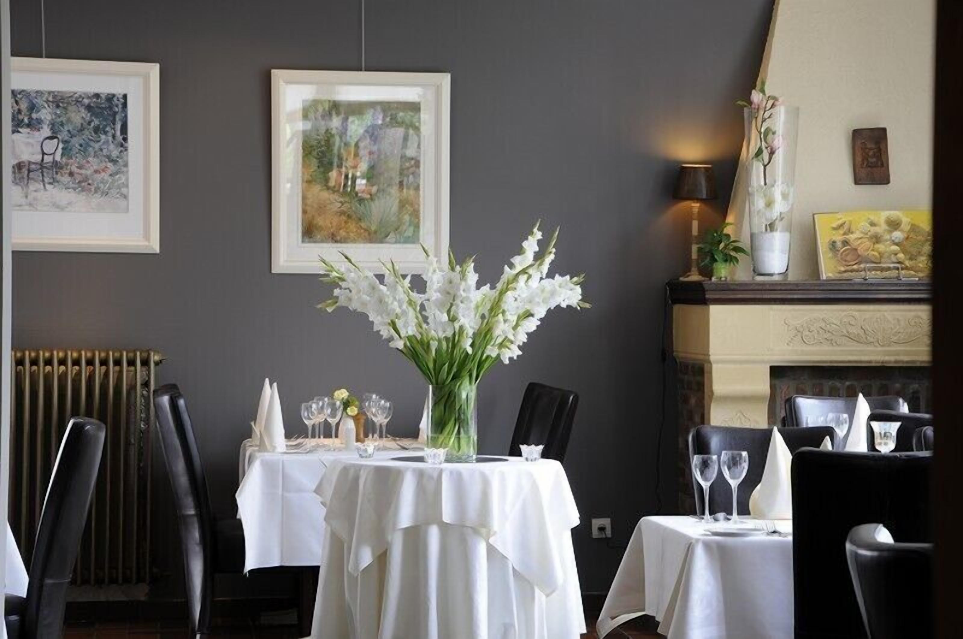A dining area with a table set for two, framed artwork on the walls, and a vase with white flowers.