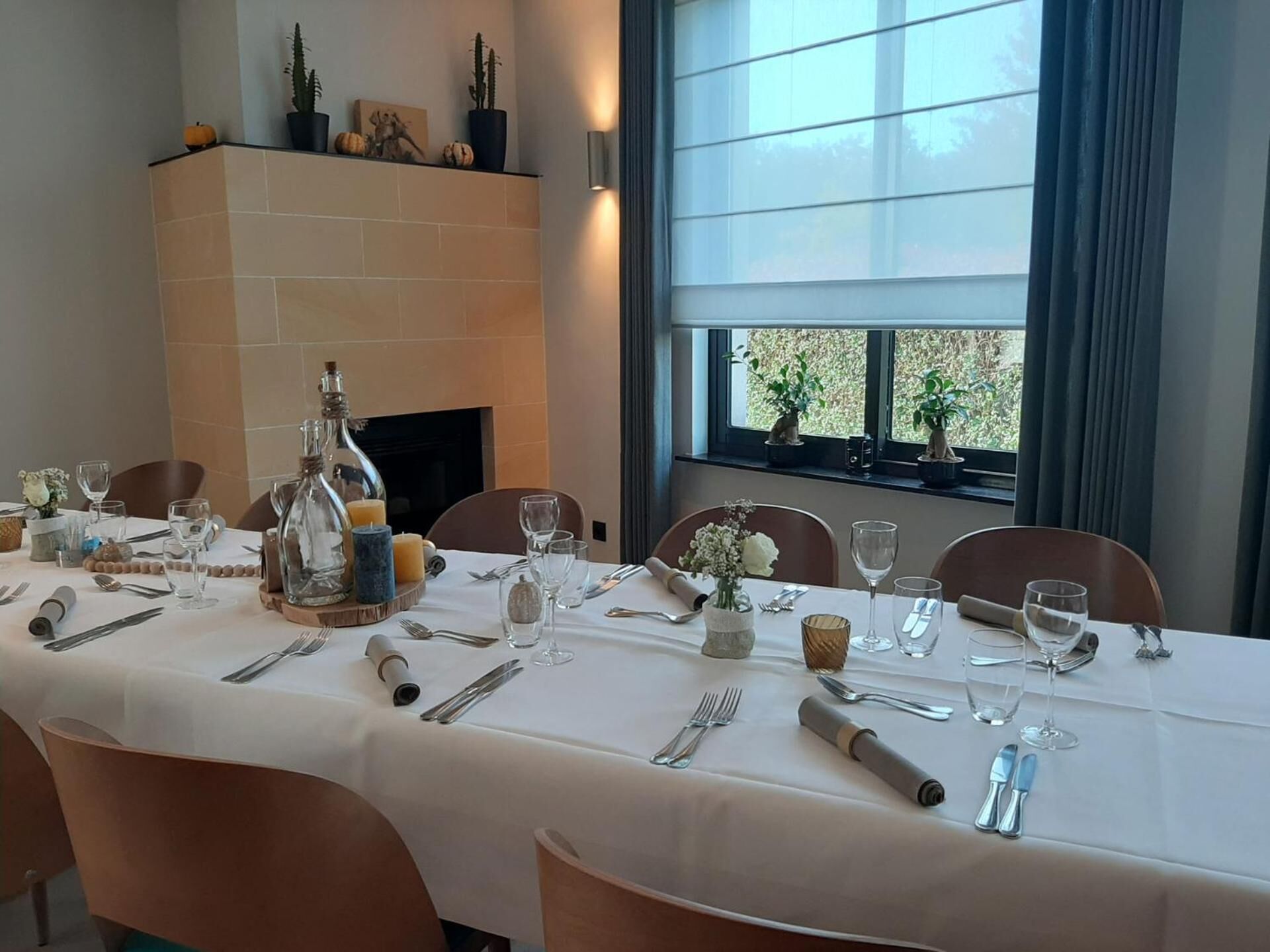 A dining table set for a meal with a fireplace in the background and a window with plants.